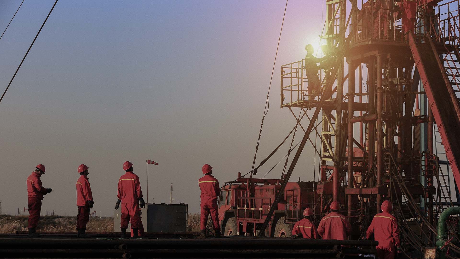 Distant, wide shot of 8 oil rig workers in red safety gear, surveying or working on an oil rig.