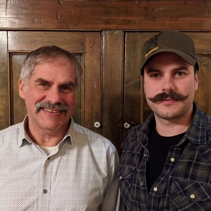 Two men with fantastic moustaches smiling to camera.