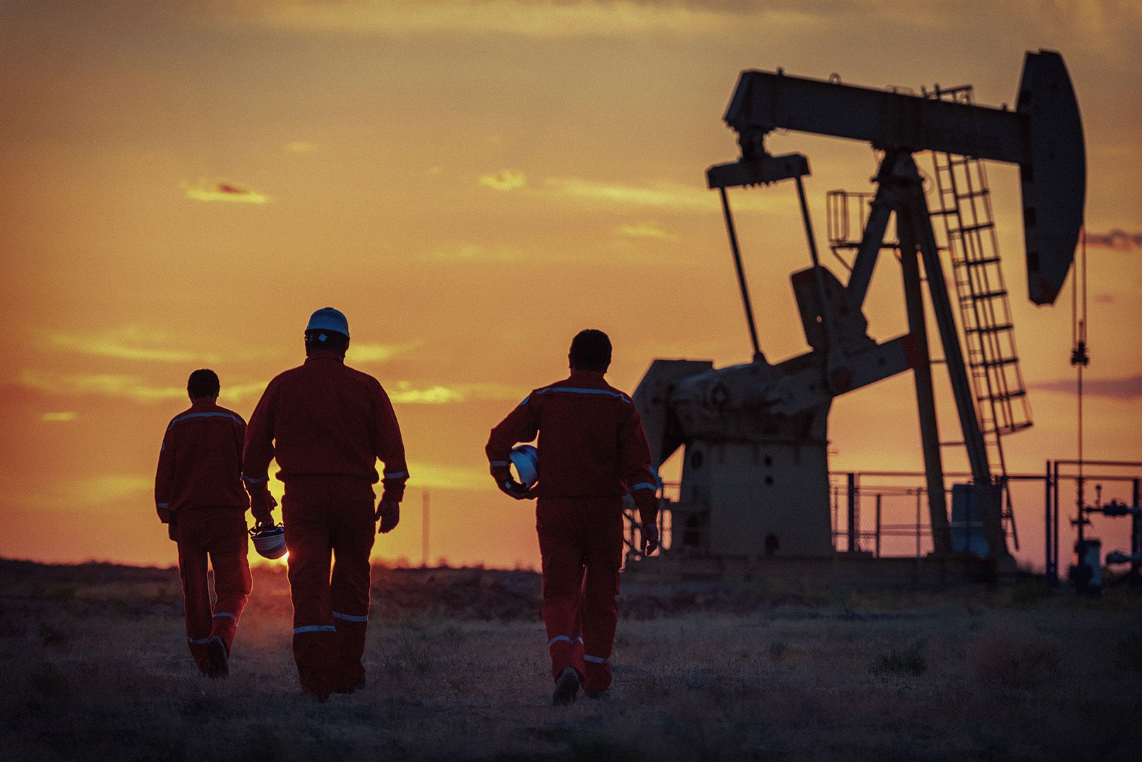 Men in construction worker uniforms and hats walking towards oil rig machinery at sunset.