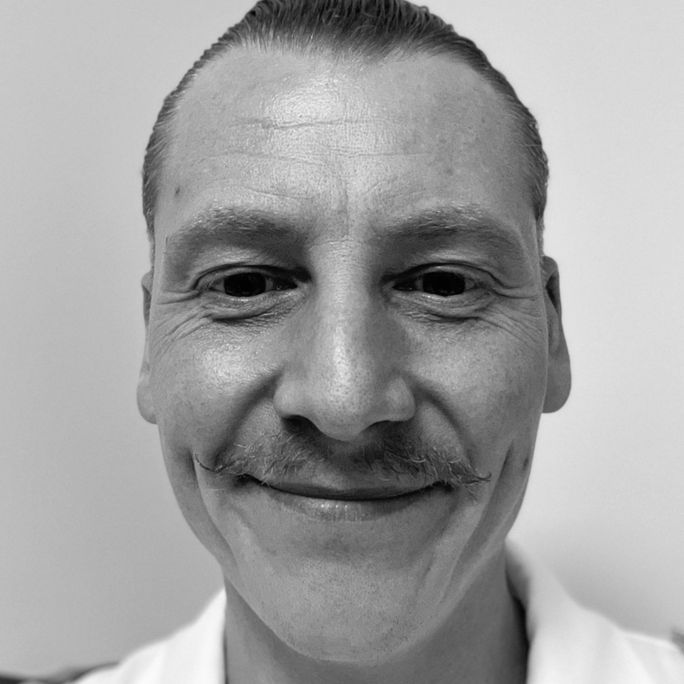 Black and white photo a man, wearing a cheeky moustache, smiling to camera.