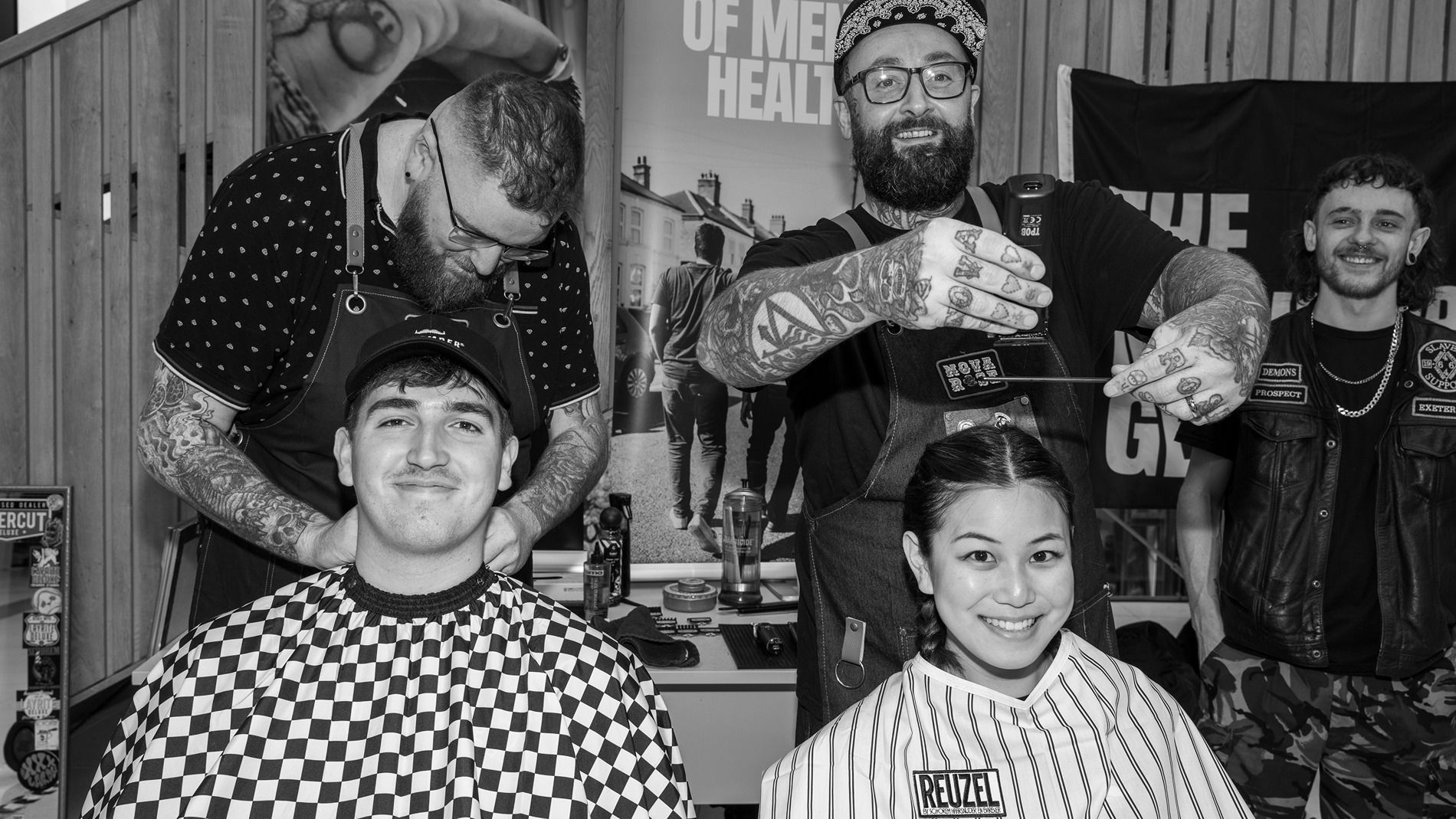 Photo of two people sitting in barber chairs, two hip-looking barbers standing behind them and about to administer haircuts.