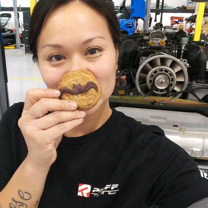 Photo of a woman looking to camera, holding a cookie with a moustache logo in front of her face.