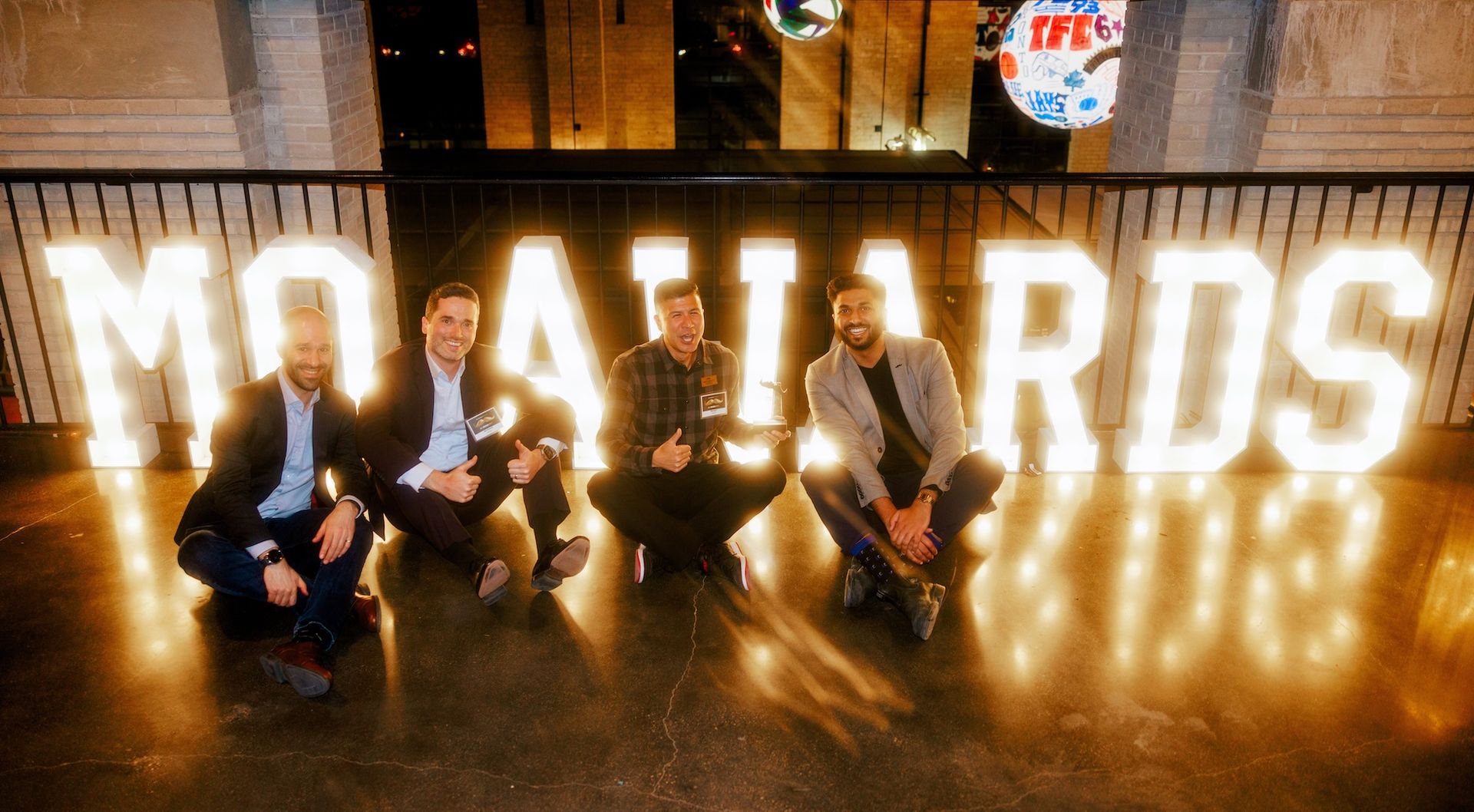 Four men sitting on the ground in front of illuminated Mo Awards signage.