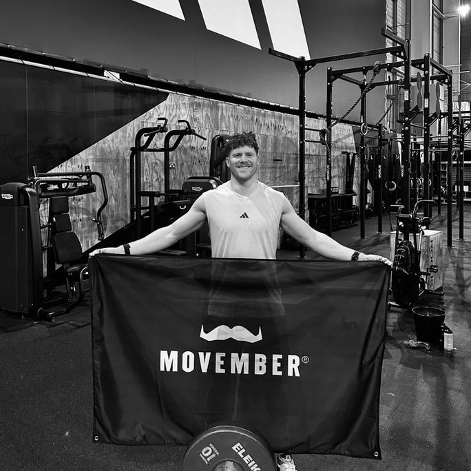 Portrait photo of a smiling man holding up a Movember-branded flag.