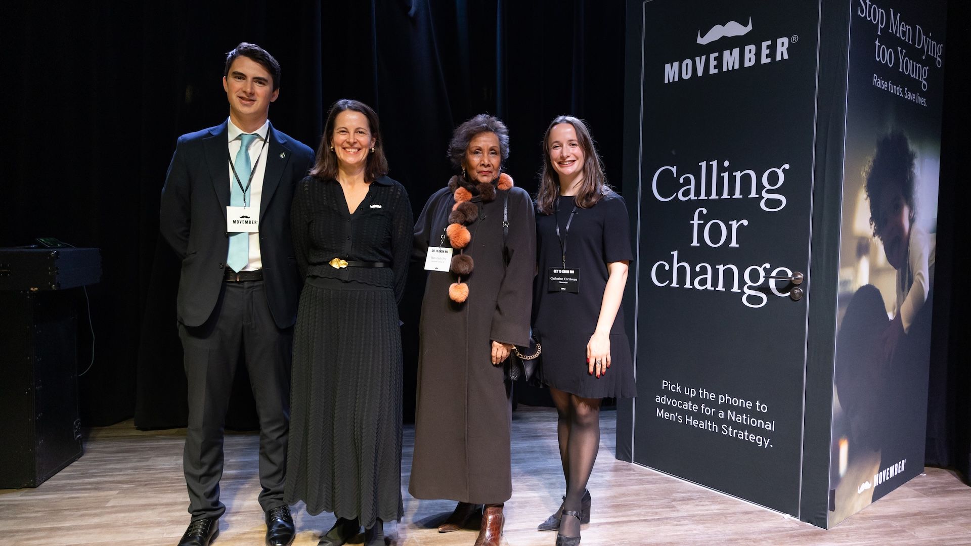 Four people pose in front of a Movember-branded phone booth at a Movember event.