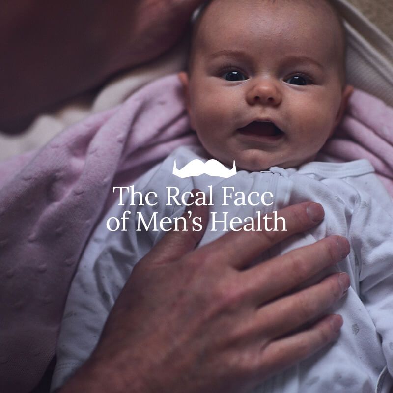 Photo of an infant looking to camera, cradled in adult hands. Superimposed text says: "The Real Face of Men's Health"
