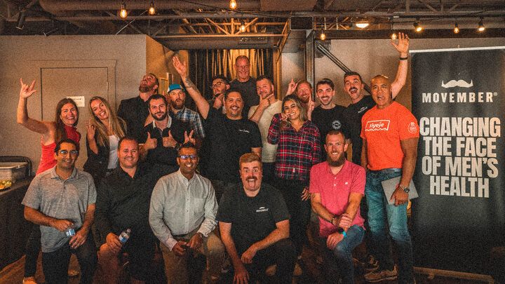 A group of smiling men and women posing in an office setting. There is a banner next to them that reads "Movember. Changing the face of Men's Health"