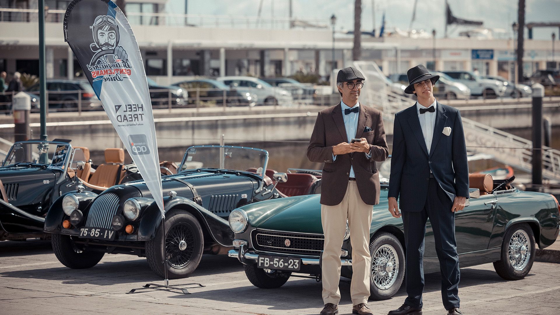 Two superbly dressed men in dapper outfits pose in front of classic cars.