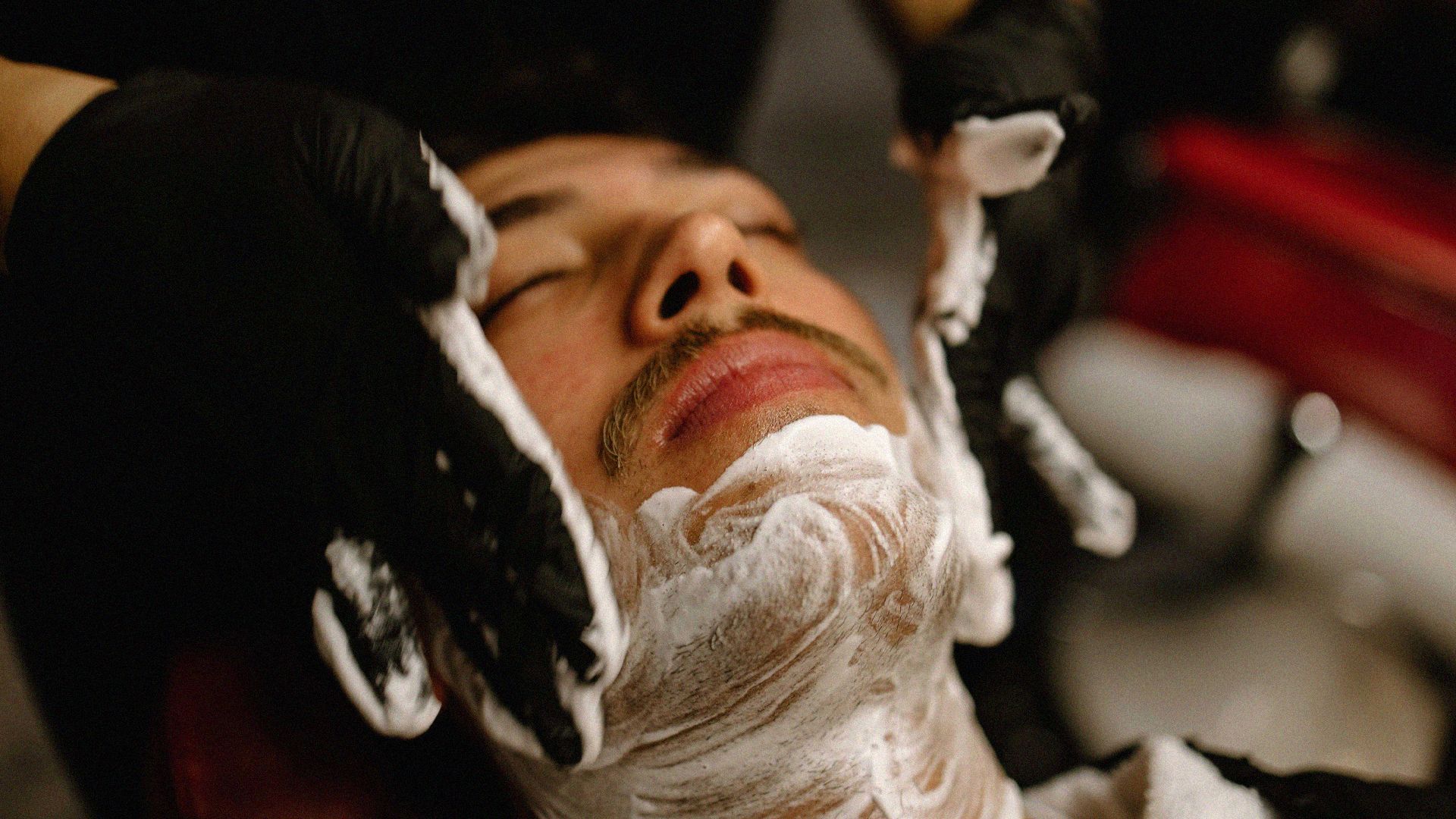 A man relaxes with his eyes closed. A barber smooths shaving cream on his face.