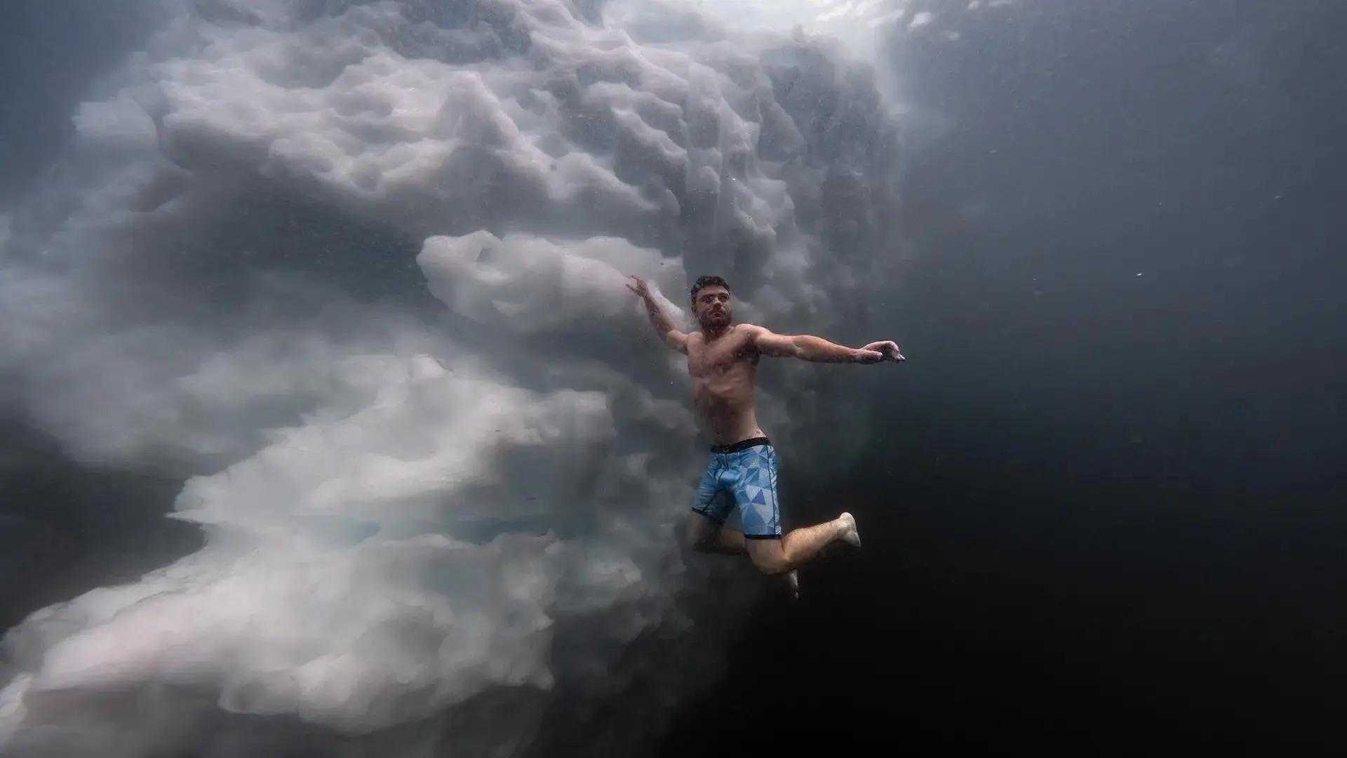 Spectacular underwater photo of man in shorts swimming below the surface next to a large block of submerged sea ice.