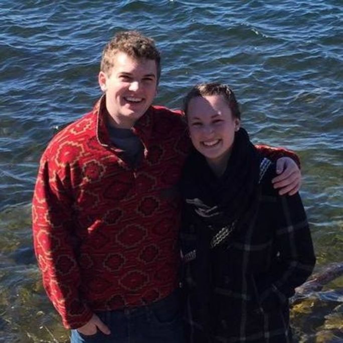 Photo of two smiling people, looking up to camera in front of a water background.
