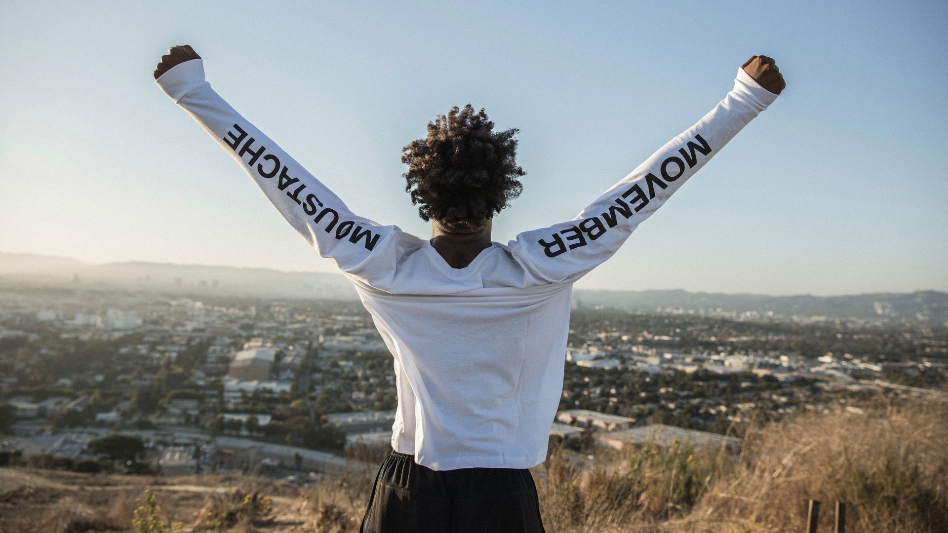 A young man stands with his arms raised over a valley. The sleeves of his shirt say "Movember" and "Moustache"