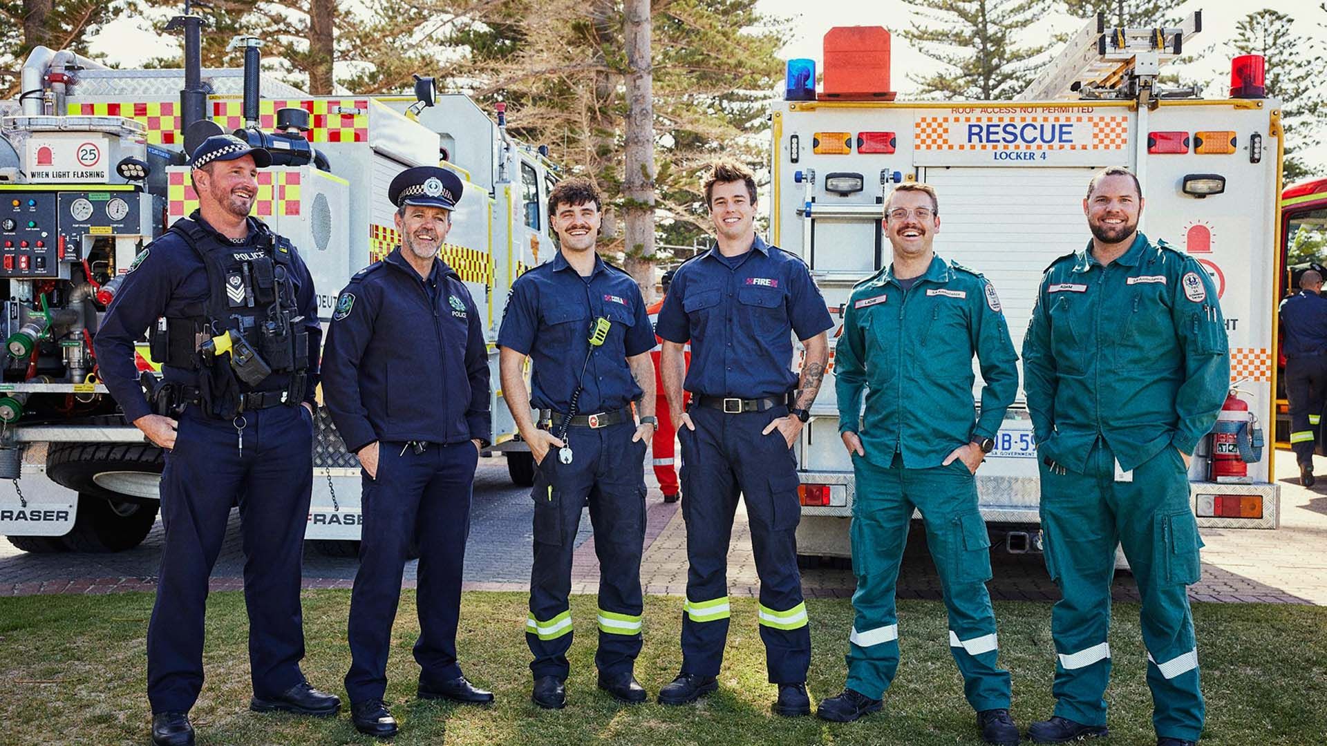 A group of emergency services men stand in a line, all in uniform, with their emergency vehicles behind them.