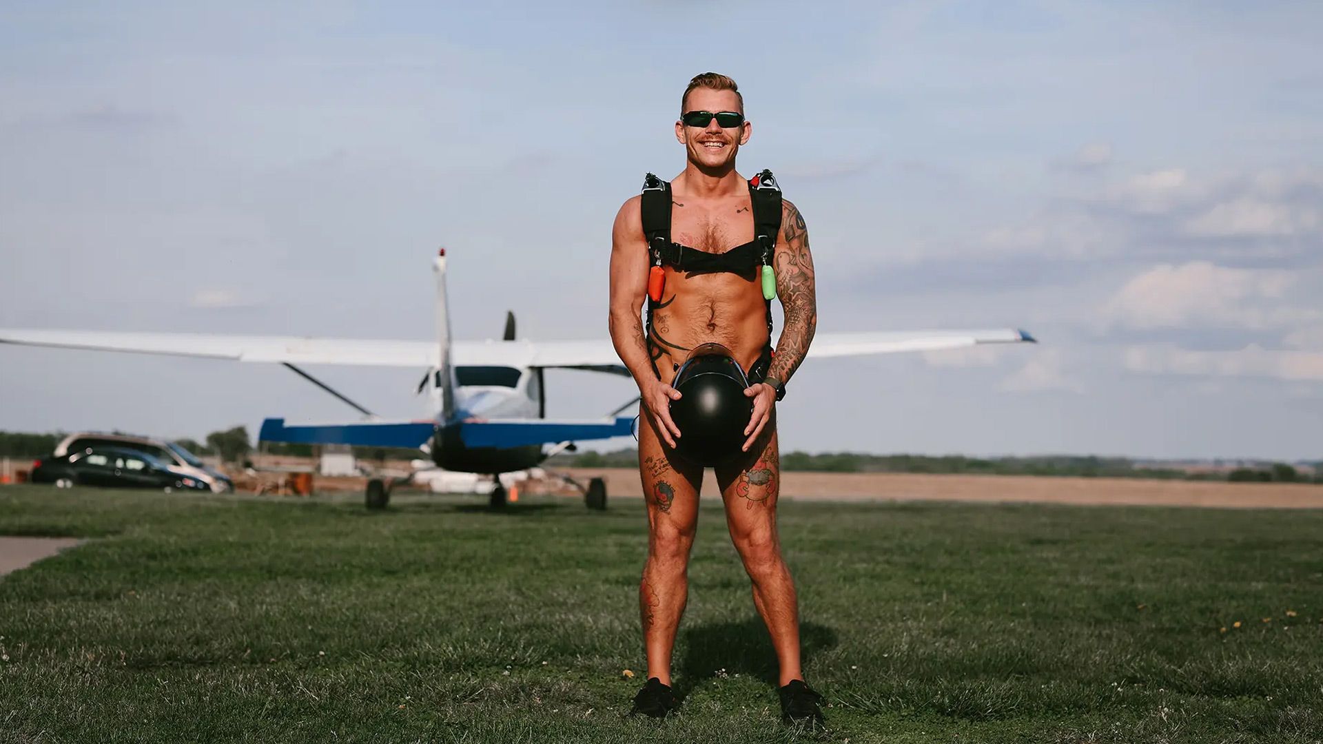 Naked skydiver smiling to camera, standing in front of aircraft.