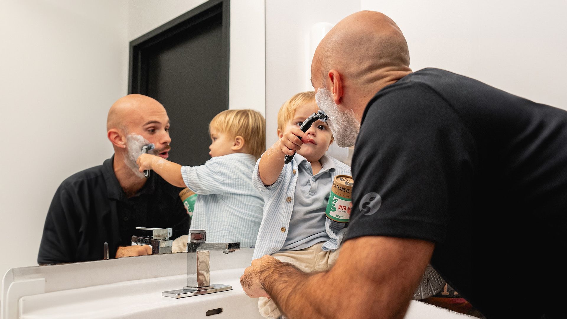  Un petit garçon assis sur le comptoir de la salle de bain aide son père à se raser le visage pour Movember