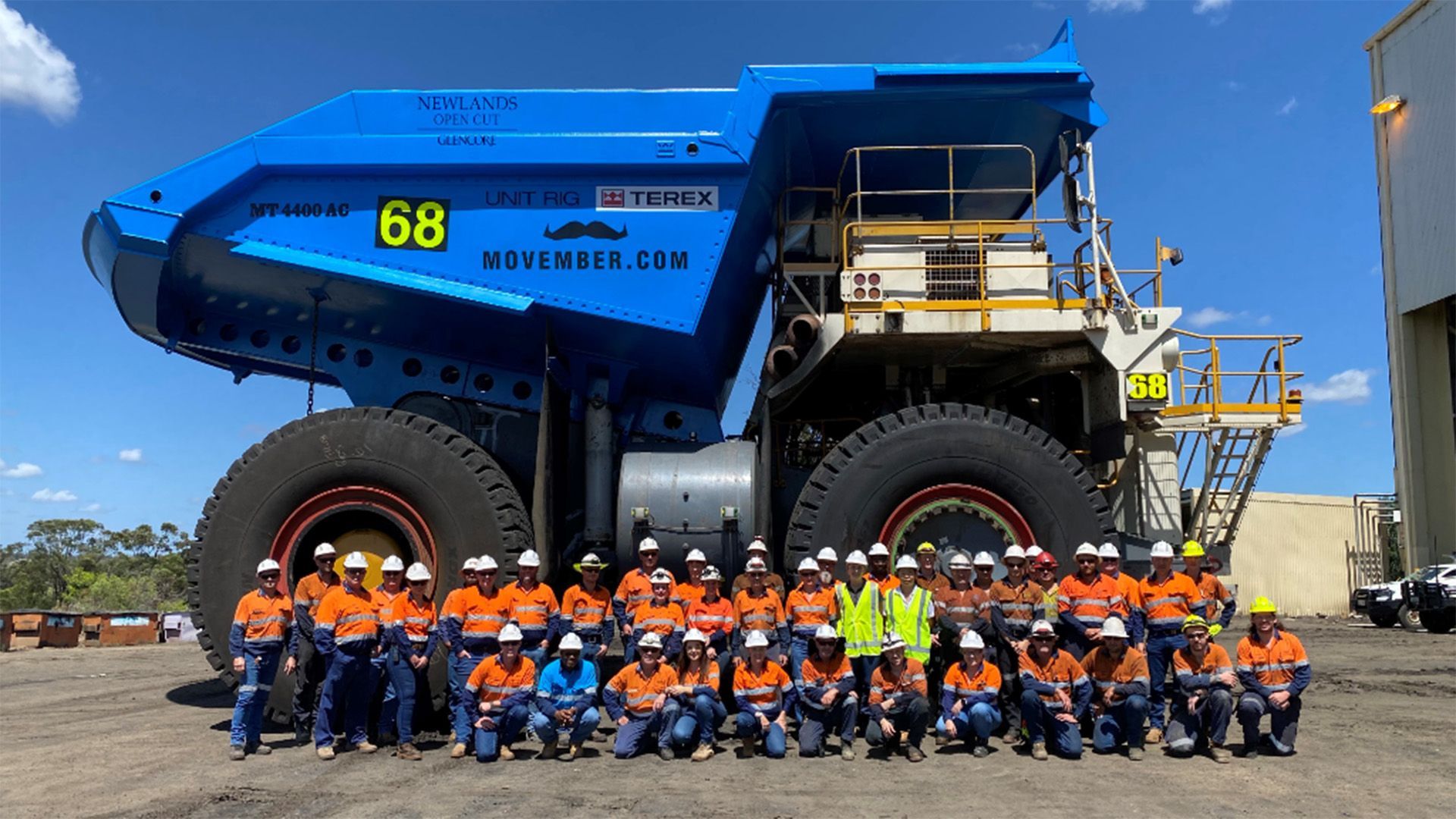 A side-view photo of a very large blue mining dump truck. A team of miners in high visibility gear pose to camera.