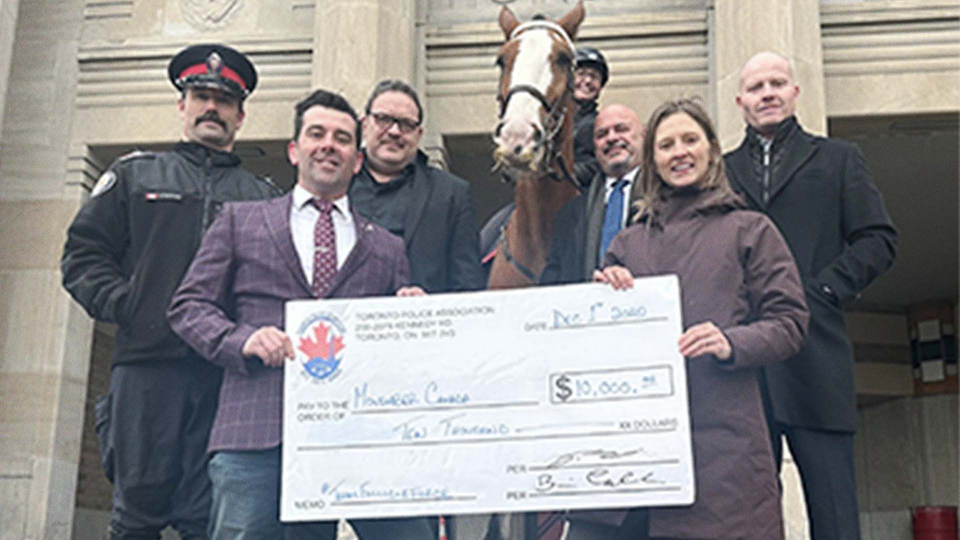Photo of a group of people holding an oversized donation cheque in front of a police horse bearing a distinct moustache.