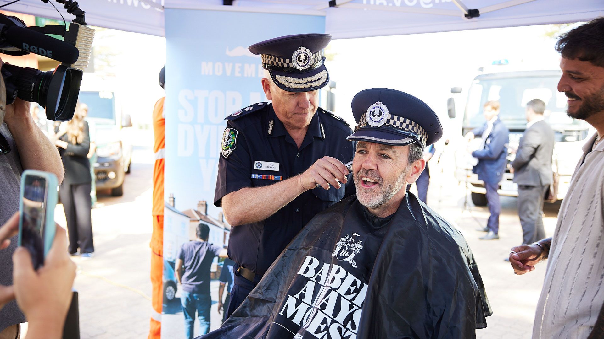 Policeman sits in barber chair with hat on, while another policeman shaves his face with clippers, both smiling.
