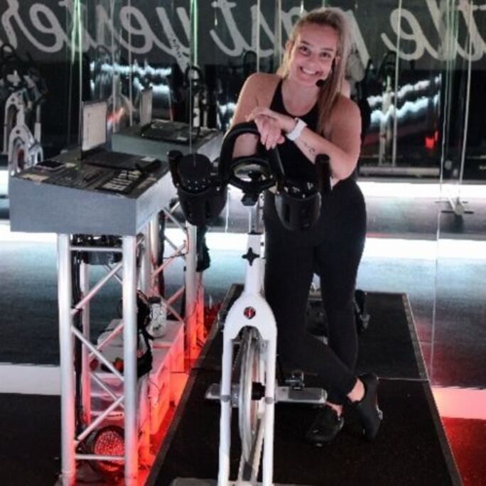 Photo of woman in sports attire, smiling to camera next to an exercise bicycle.