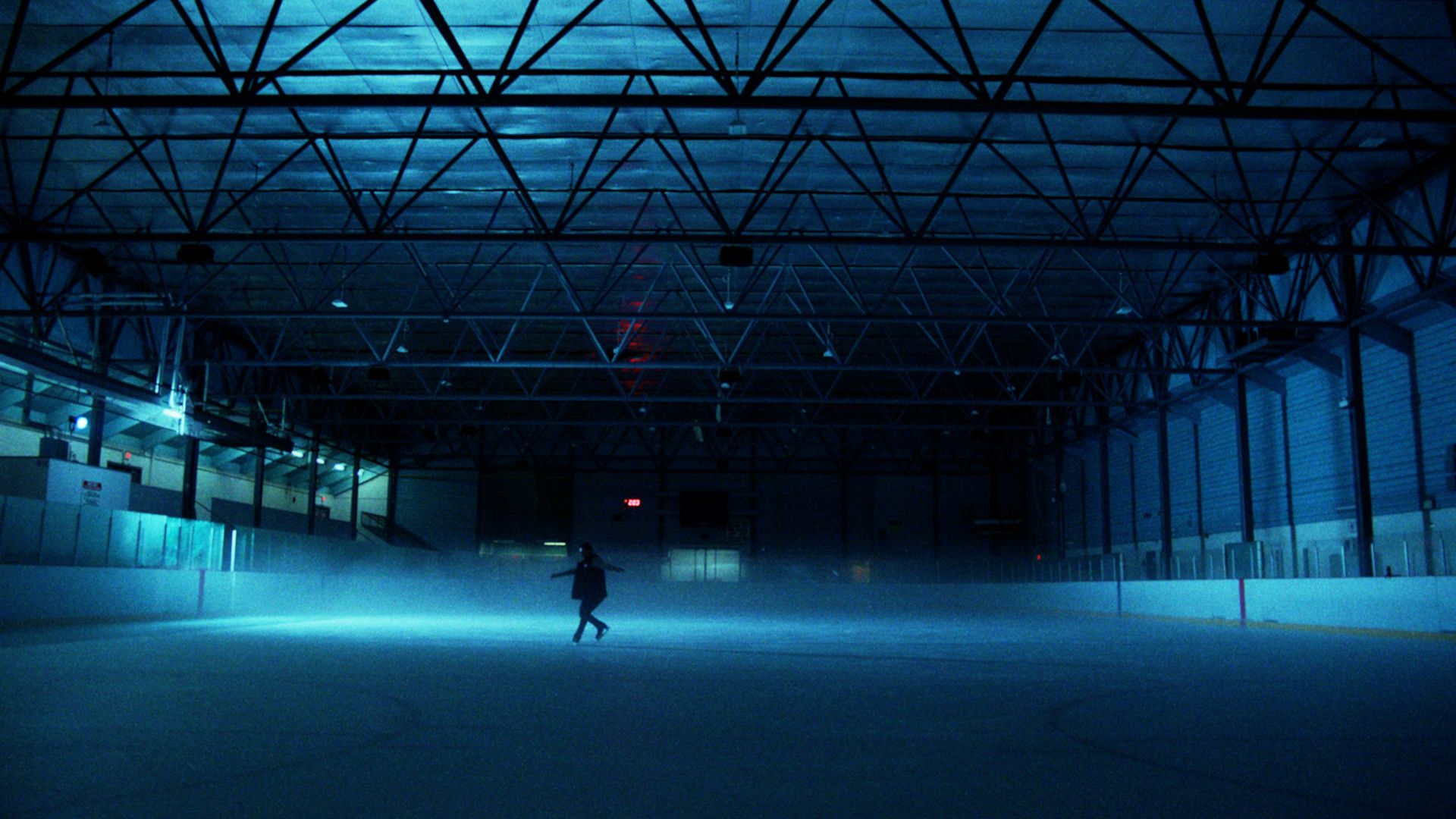 Wide photo of a dimly lit ice skating rink, with a solitary silhouetted figure skating in the middle ground.