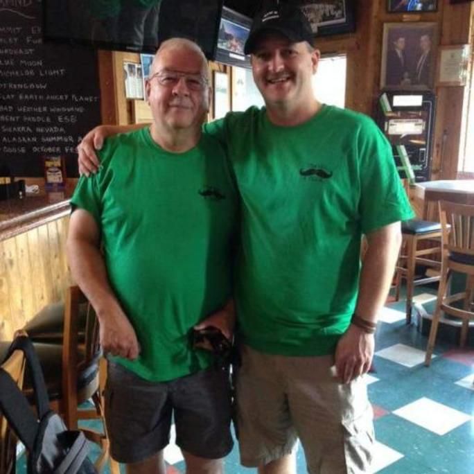 Photo of two men wearing green Movember-branded shirts in a bar, posing to camera.