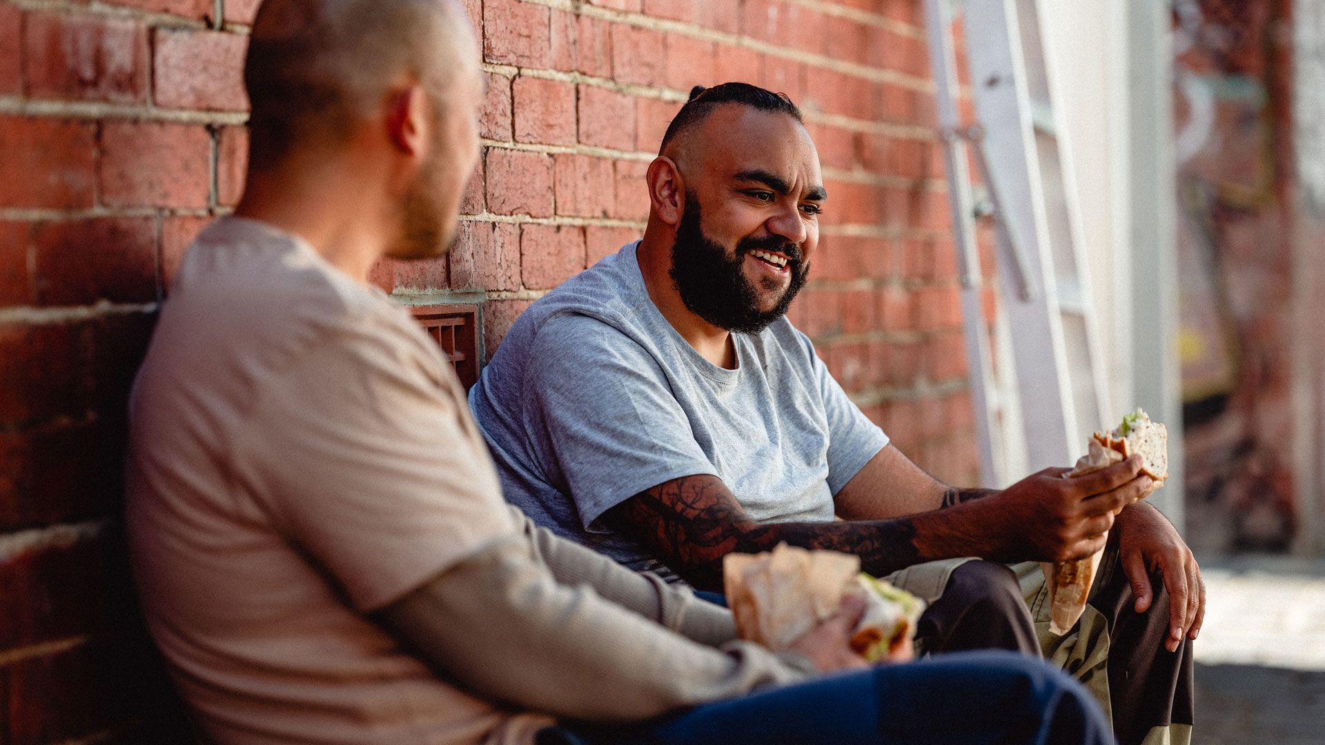 Two smiling men, sitting in a laneway while enjoying their lunchbreak.