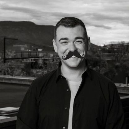 Black and white photo of man donning what appears to be a cartoon moustache, smiling to camera.
